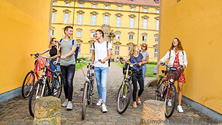 Eine Gruppe von Studierenden schiebt ihre Fahrräder durch das Haupttor des Schlosses.