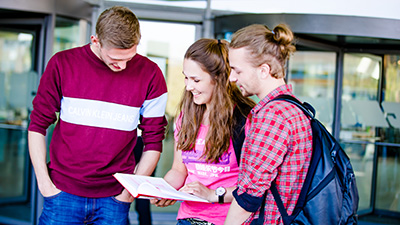 Drei Studierende vor dem "Frosch"-Gebäude auf dem Campus Westerberg schauen gemeinsam in ein Buch.