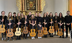 15 Mitglieder des Gitarrenensembles stehen für ein Gruppenfoto vor einem Altarbild. Sie halten ihre Gitarren vor sich. Ganz rechts steht Leiter Tim Sandkämper.