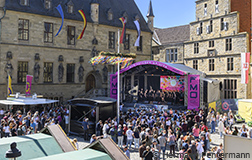 Eine große Menschenmenge versammelt sich auf einem Platz vor einer Bühne, auf der ein Chor auftritt. Die Bühne ist mit bunten Plakaten und Lichtern dekoriert. Im Hintergrund sind historische Gebäude zu sehen. Es ist helles Wetter und die Atmosphäre ist festlich.