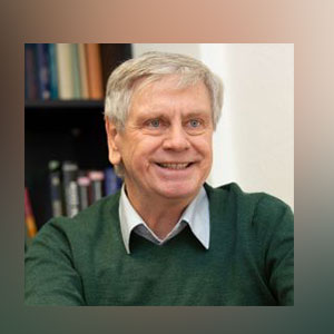 Ein Mann mit grauen Haaren und grünem Pullover sitzt vor einer Bücherwand und schaut  lächelnd Richtung Kamera